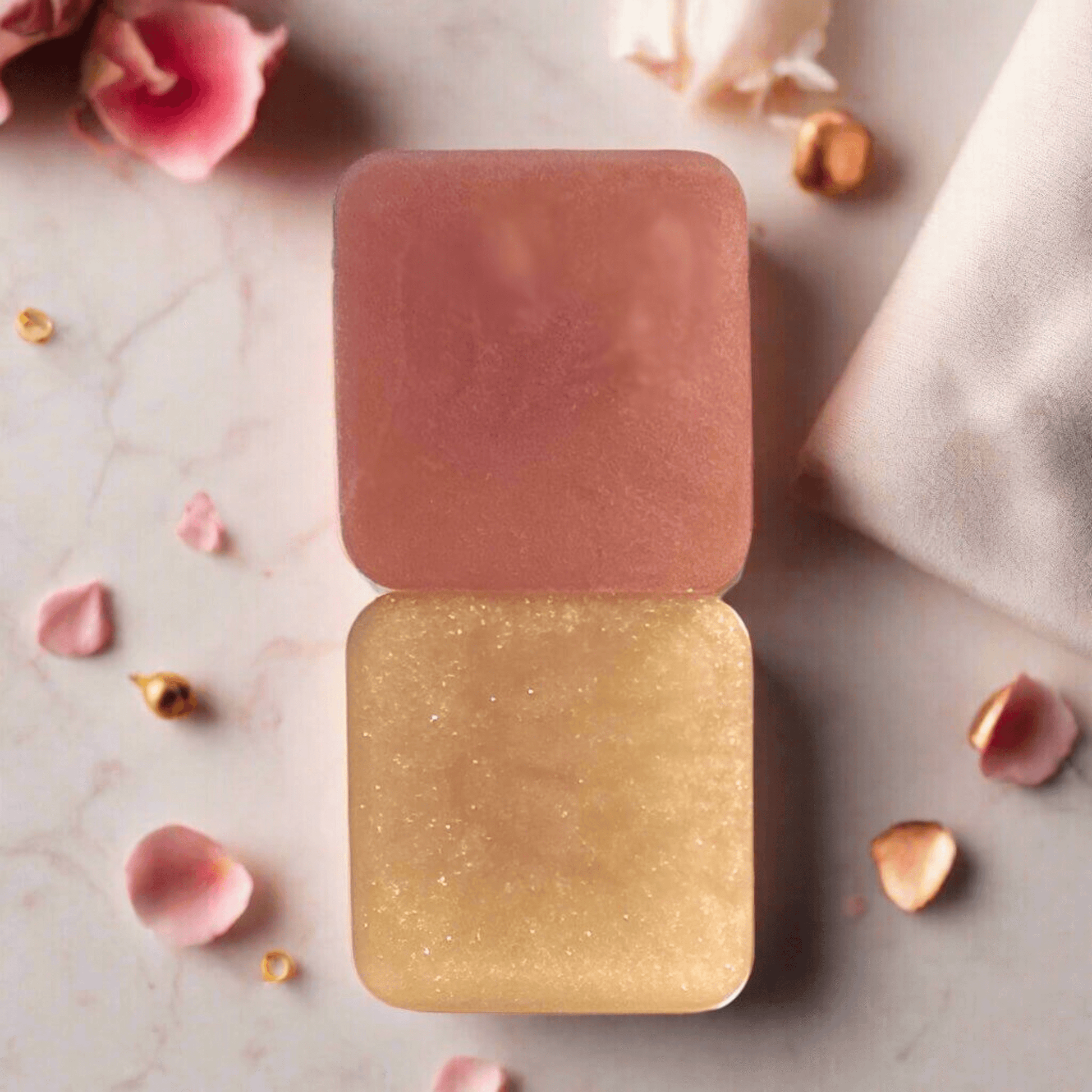 Two bars of Rosegold Rhapsody laying on a marble counter surrounded by rose petals, cashmere scarf, and rosegold pieces. 