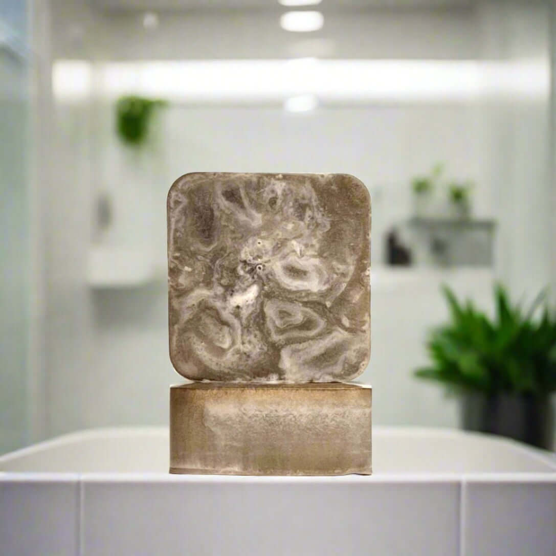 Two Tobacco + Mint Timbre soap bars sitting on the edge of a white bathroom sink with green plants in the background.